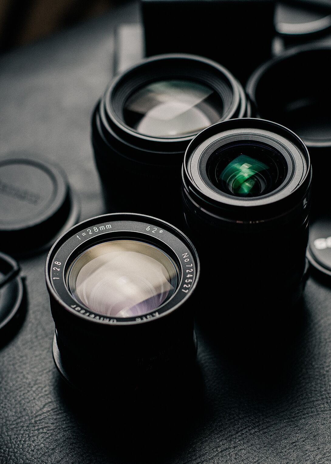 Several camera lenses lie side by side on a dark surface, some with color-accentuated glass reflections, depicted in a detailed close-up.