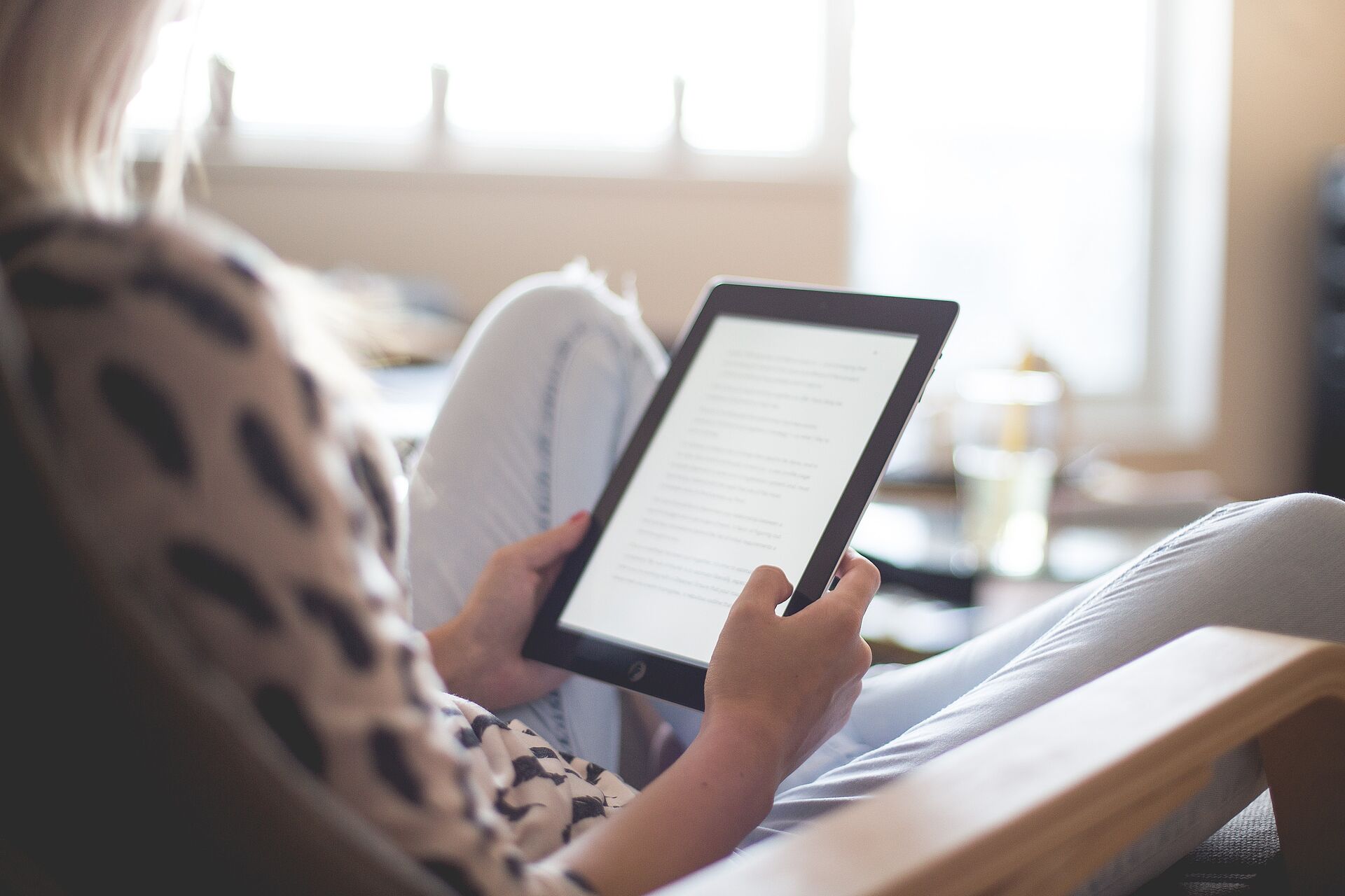 Person hält ein Tablet und liest einen Text, entspannt sitzend in einem hellen Wohn- oder Leseumfeld mit unscharfem Hintergrund.