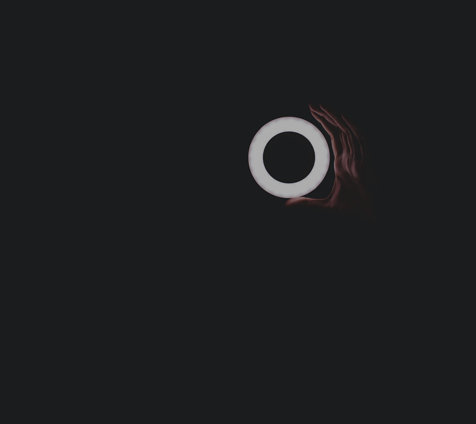 A glowing, circular ring of light is held by a human hand against a dark background, creating an abstract, minimalist lighting mood.